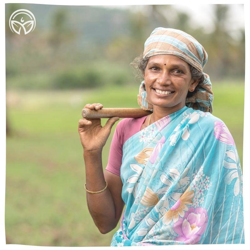 Smiling woman in a floral blue saree and headscarf, holding a tool on her shoulder. She stands in a green field, conveying a sense of joy and rural life.