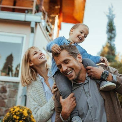A joyful family moment outdoors, with a father carrying a smiling child on his shoulders and the mother looking on happily. The background features a modern house and trees.