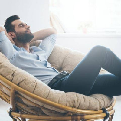 A man relaxes in a plush round chair with his eyes closed and arms behind his head. He wears a light blue shirt and jeans, exuding a serene and content mood.