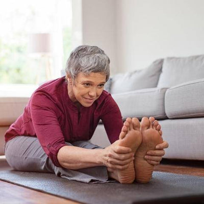 Elderly person in a red shirt and gray pants performs a seated forward bend on a yoga mat in a cozy living room. The mood is focused and calm.