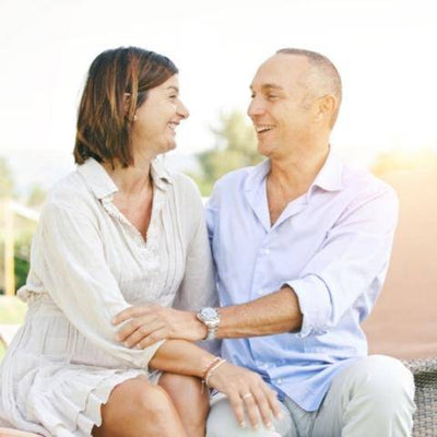 A smiling couple sits outdoors on a sunny day, embracing and gazing at each other warmly. The background is softly blurred, conveying a sense of happiness and connection.