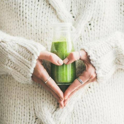 Person in a cozy white knit sweater holding a small bottle of green smoothie with hands forming a heart shape. Calm and comforting atmosphere.
