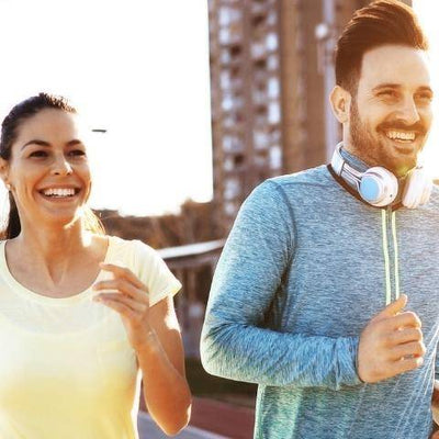 A man and woman jogging outdoors, smiling brightly. She wears a yellow shirt, and he has a blue top with headphones around his neck, conveying joy and vitality.
