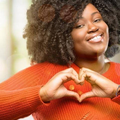 Smiling woman with curly hair wearing an orange sweater forms a heart shape with her hands. Background is softly blurred, conveying warmth and positivity.