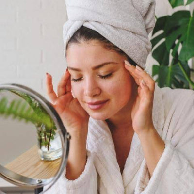 A woman in a white robe with a towel on her head gently touches her face, smiling contently. She sits by a mirror and a plant, conveying relaxation.